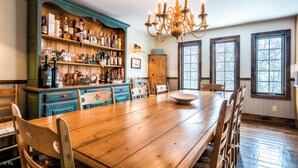 Une table à manger en bois avec des chaises, un coin bar avec des étagères remplies de bouteilles et un lustre.