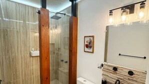 A bathroom with a glass-enclosed shower, a white toilet, a wooden vanity with a stone backsplash, and a mirror with light fixtures.