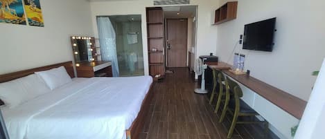 A hotel room with a large bed, a desk with a chair, a TV, and a bathroom visible through a glass door.