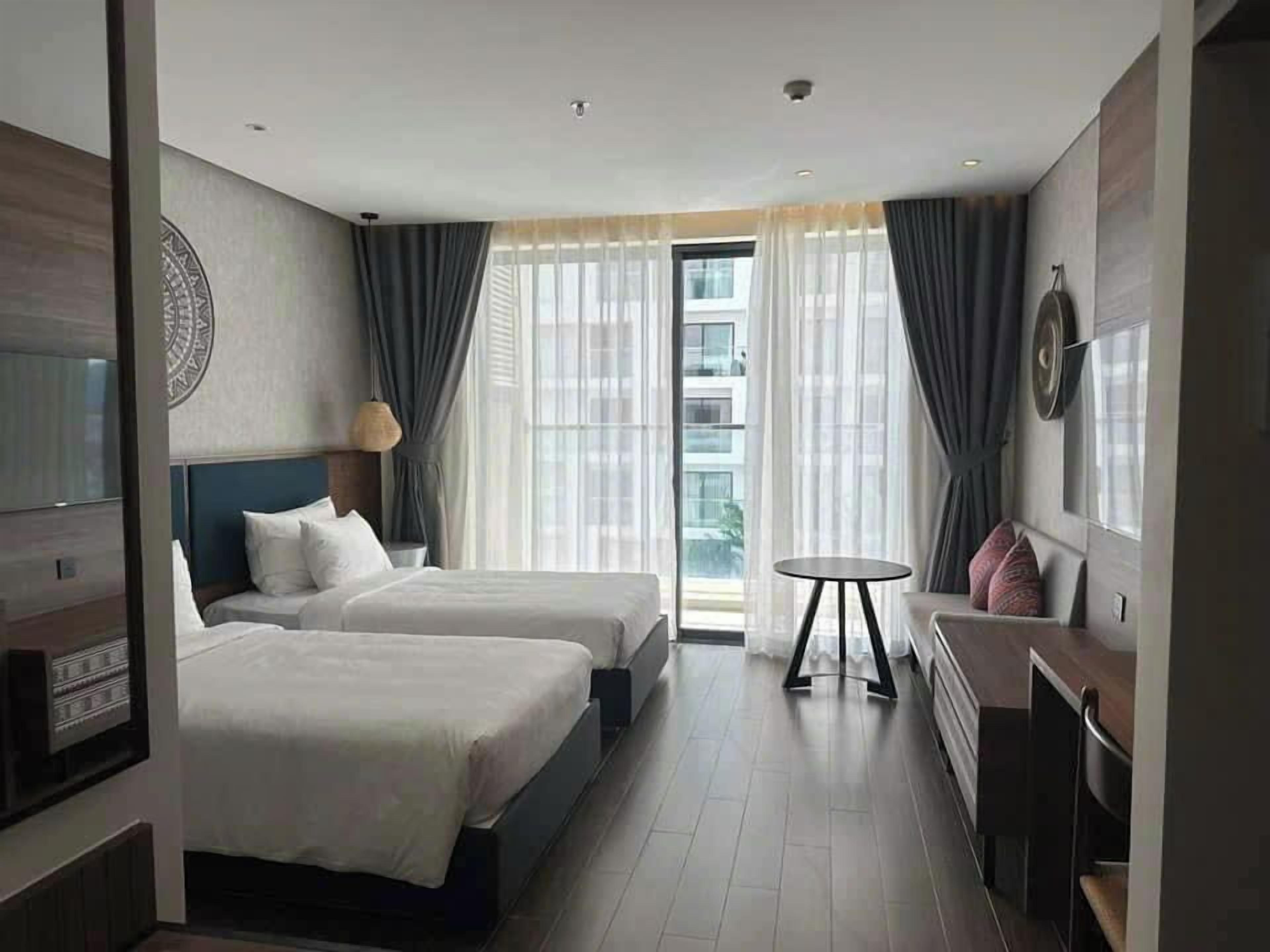 Una habitación de hotel con dos camas, una mesita, un televisor y una ventana con cortinas.