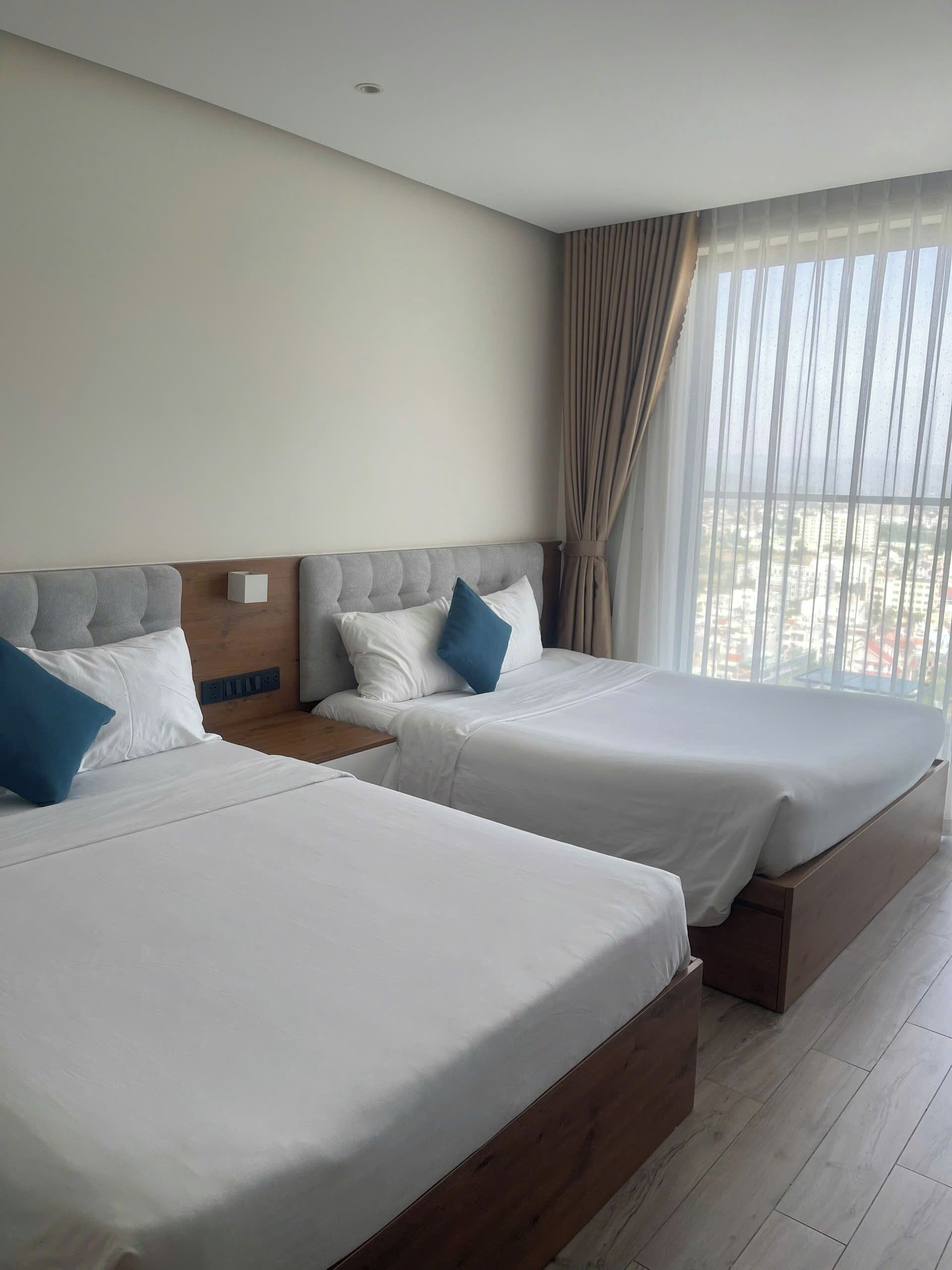 Habitación de hotel con dos camas, cabecero de madera, mesita de noche y ventana con cortinas.