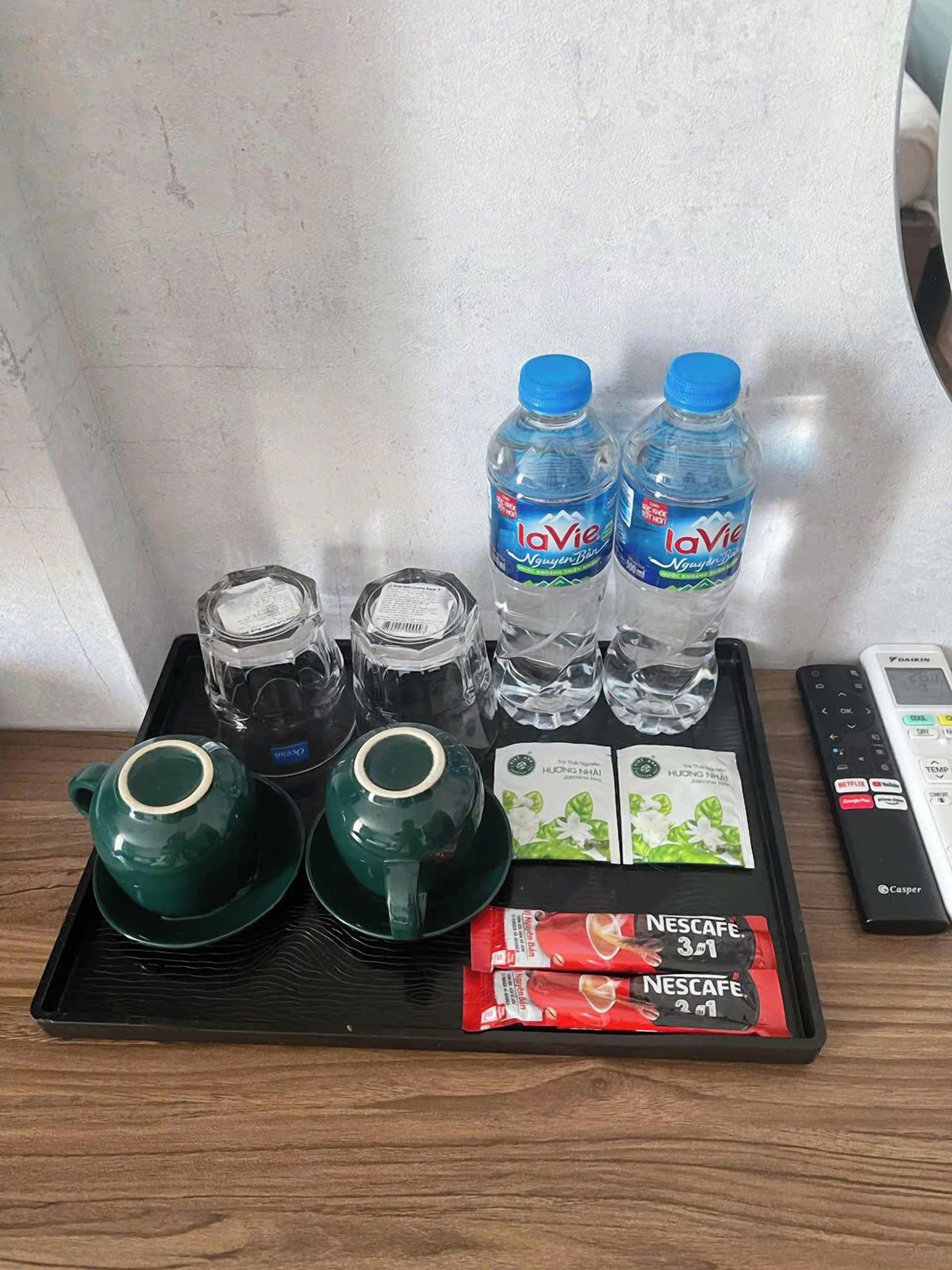 A tray with two LaVie water bottles, two glass tumblers, two green teapots, two packets of Nescafe 3 in 1, and a remote control.