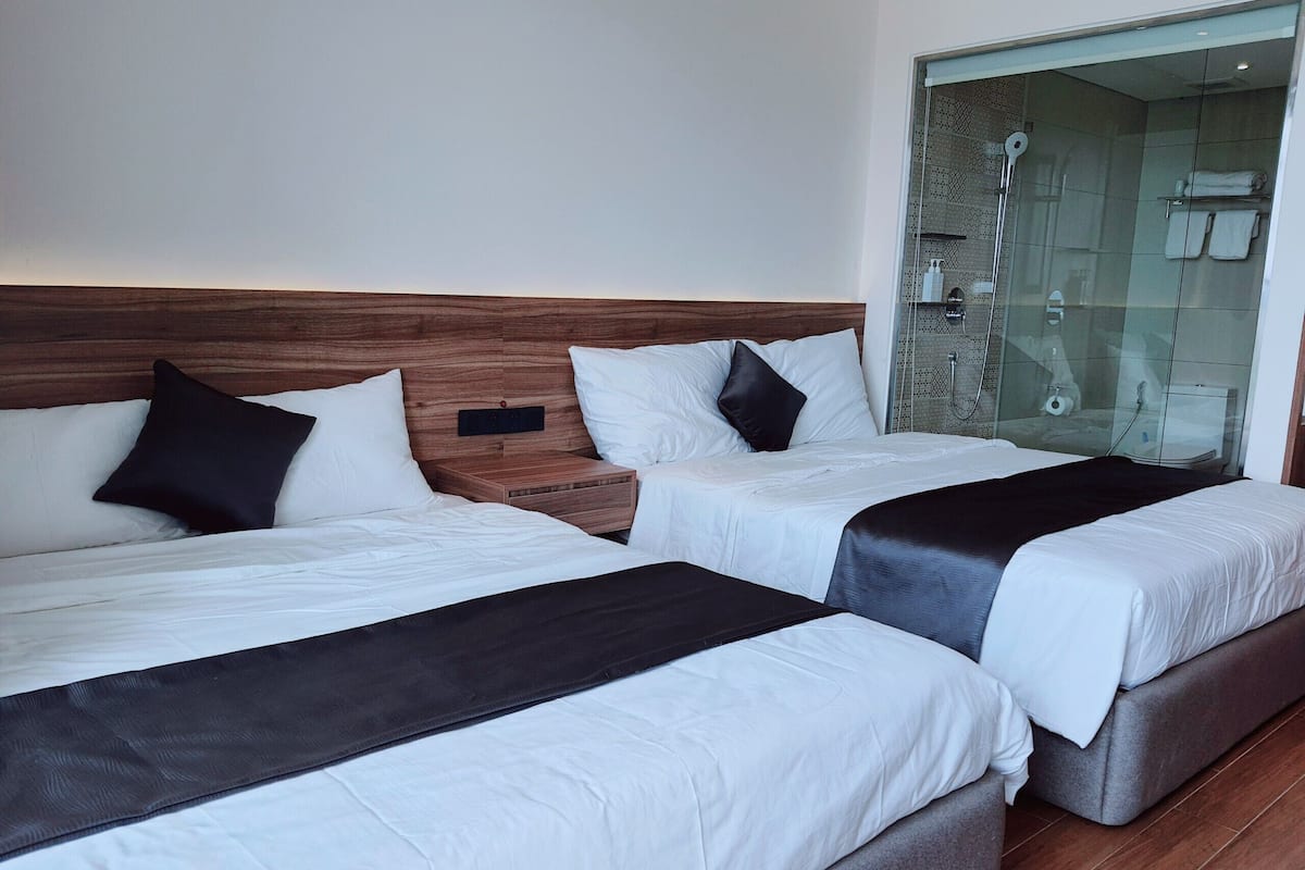 Habitación de hotel con dos camas, cabeceros de madera, ropa de cama blanca y zona de ducha con mampara de cristal.