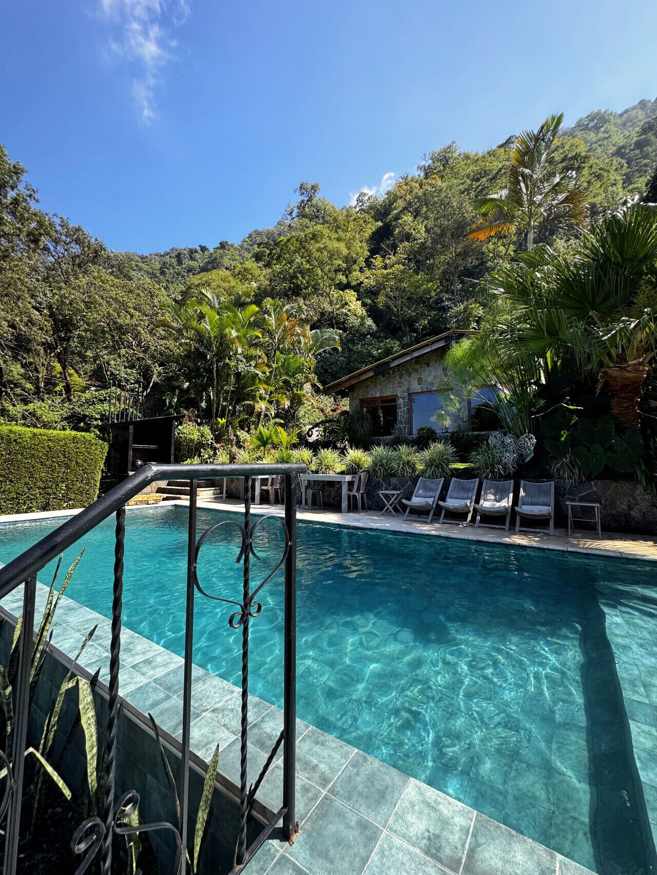 Ein klares Schwimmbecken mit einem Steinhaus im Hintergrund, umgeben von üppigem Grün und Bäumen.