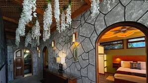 Ein Korridor mit Steinwänden, an dem weiße Blüten hängen, eine hölzerne Bank und ein Bett mit Kopfteil und Kissen.