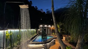 Ein Schwimmbecken mit Wasserfall, umgeben von tropischen Pflanzen und einem Steinweg, der zu einer überdachten Sitzgelegenheit führt.