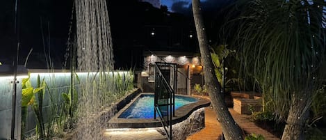 A swimming pool with a waterfall feature, surrounded by tropical plants and a stone pathway leading to a covered seating area.