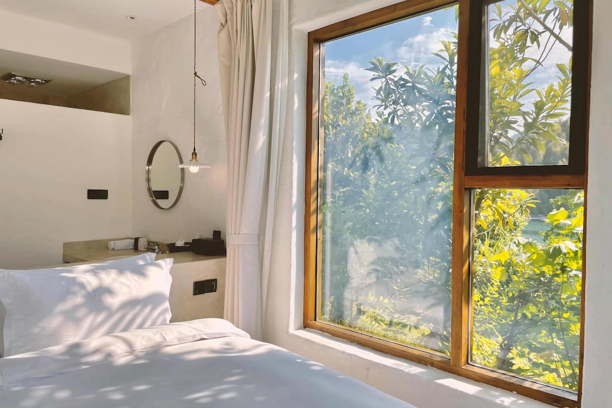 Ein Schlafzimmer mit großem Fenster, einem Bett, einem Nachttisch, einem Spiegel und einer Pendelleuchte.