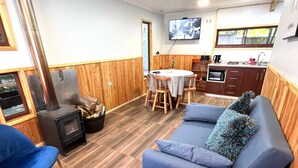A cozy living area with a sofa, chairs, a small table, a TV, and a kitchen area with wooden cabinets and a stove. - Cabañas El Nativo 2 - Licán Ray (Villarrica)
