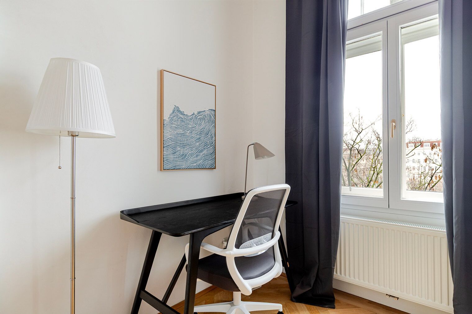 Ett rum med ett svart skrivbord, en vit stol, en golvlampa, ett inramat konstverk och ett fönster med gardiner.