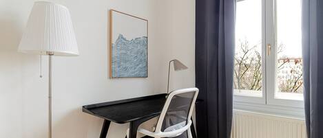 Ett rum med ett svart skrivbord, en vit stol, en golvlampa, ett inramat konstverk och ett fönster med gardiner.