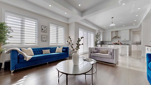 A modern living room with a blue sofa, a grey sofa, and a round glass table. There are large windows with blinds, a kitchen in the background, and a ceiling with recessed lighting. - A cozy getaway for a quiet relaxing spell. (Vaughan)