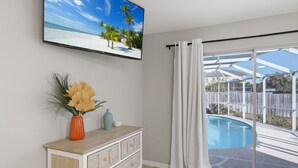 Een kamer met een grote televisie waarop een strand te zien is, een houten dressoir, een vaas met gedroogde bloemen en een glazen schuifdeur naar een zwembad.
