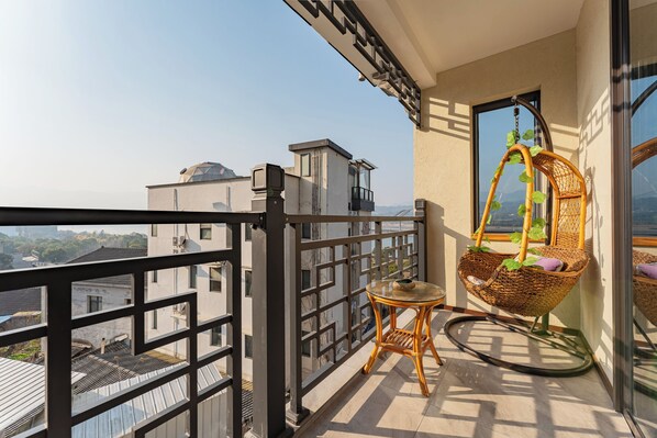 A balcony with a wicker chair, a small table, and a view of buildings and greenery.
