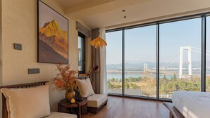 A hotel room with a large window, a bed, a sofa, a small table, and a painting of a mountain.