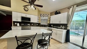 A modern kitchen with white cabinets, a large island with two bar stools, a ceiling fan, and a view of a swimming pool through large sliding glass doors.