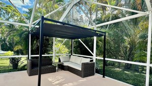 A covered outdoor seating area with a swing, surrounded by a metal frame and glass walls, overlooking a lush garden.