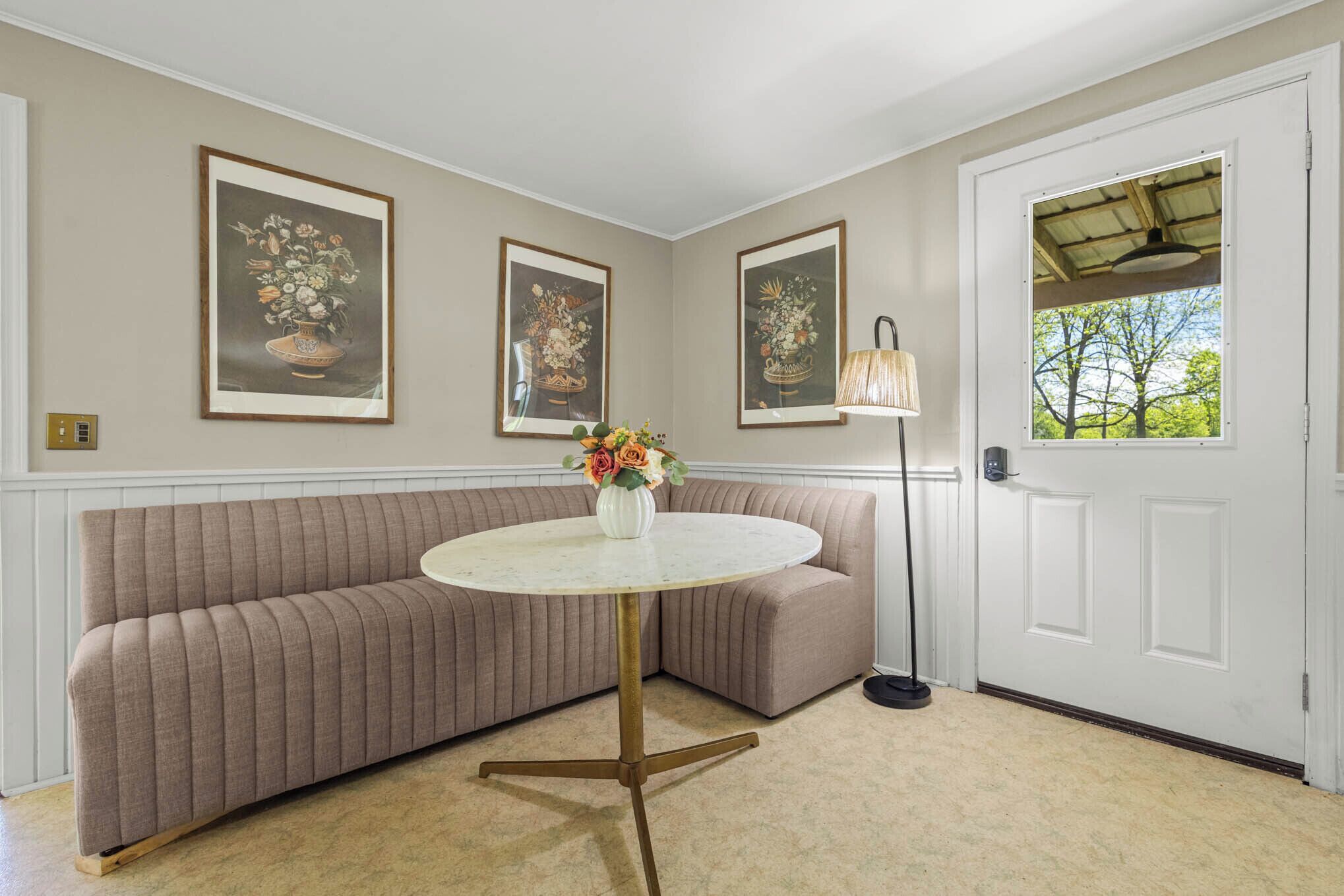 A room with a beige sofa, a round table with a vase of flowers, framed floral paintings on the wall, a floor lamp, and a door with a view of trees.