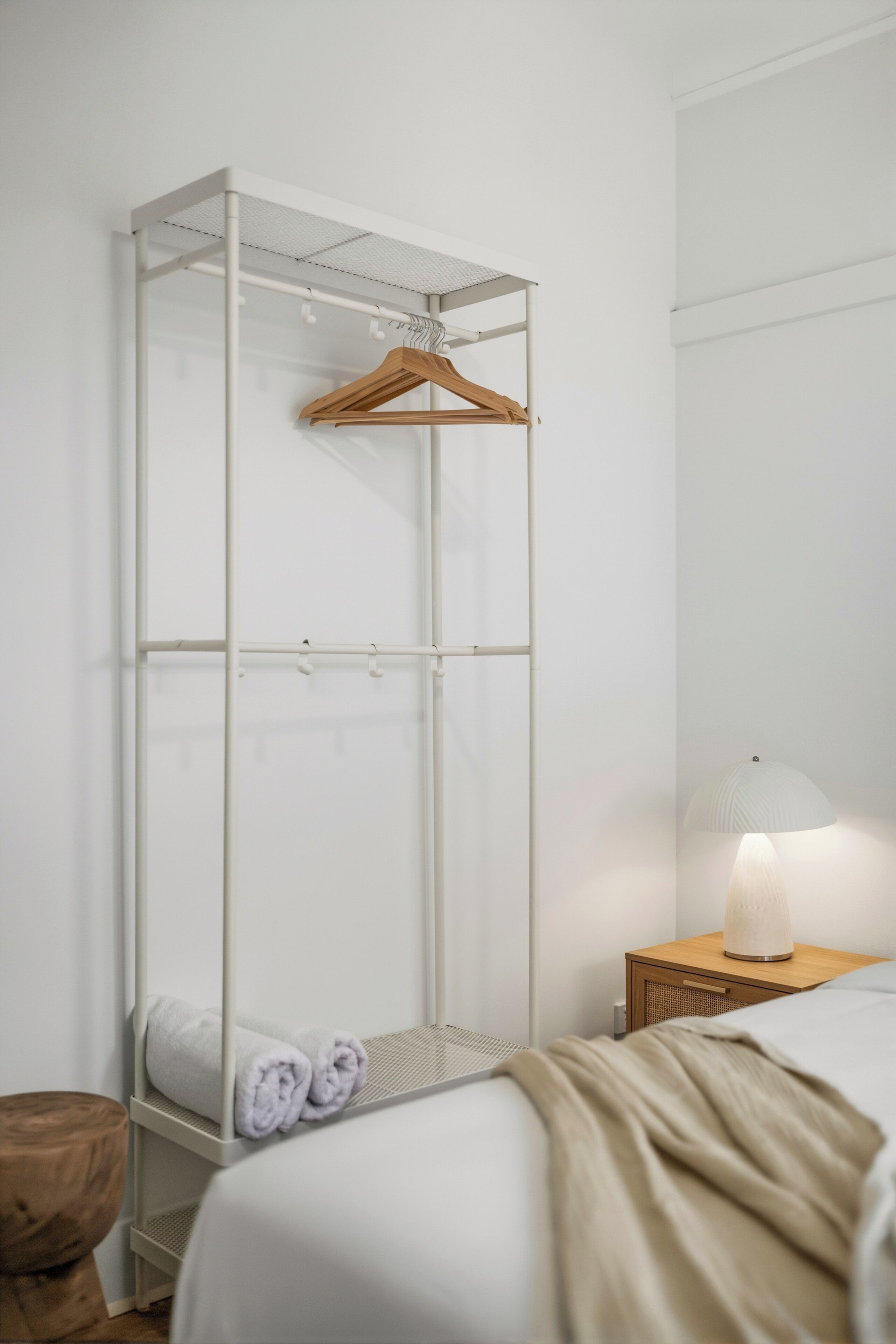 Un dormitorio con cama, una mesita de noche con lámpara, una mesita auxiliar de madera, un armario blanco con perchero de madera y una toalla enrollada en el suelo.