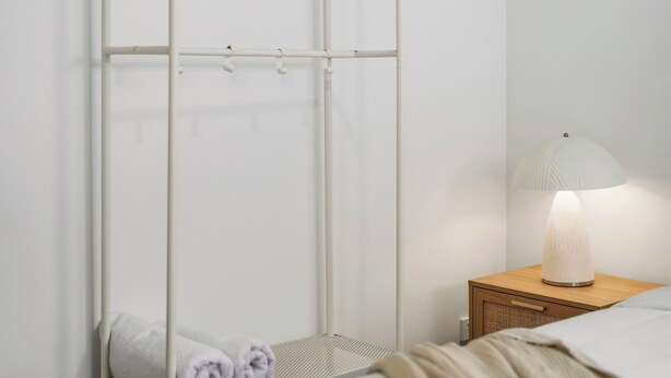 Une chambre à coucher avec un lit, une table de chevet avec une lampe, une petite table en bois, une armoire blanche avec un cintre en bois, et une serviette enroulée sur le sol.