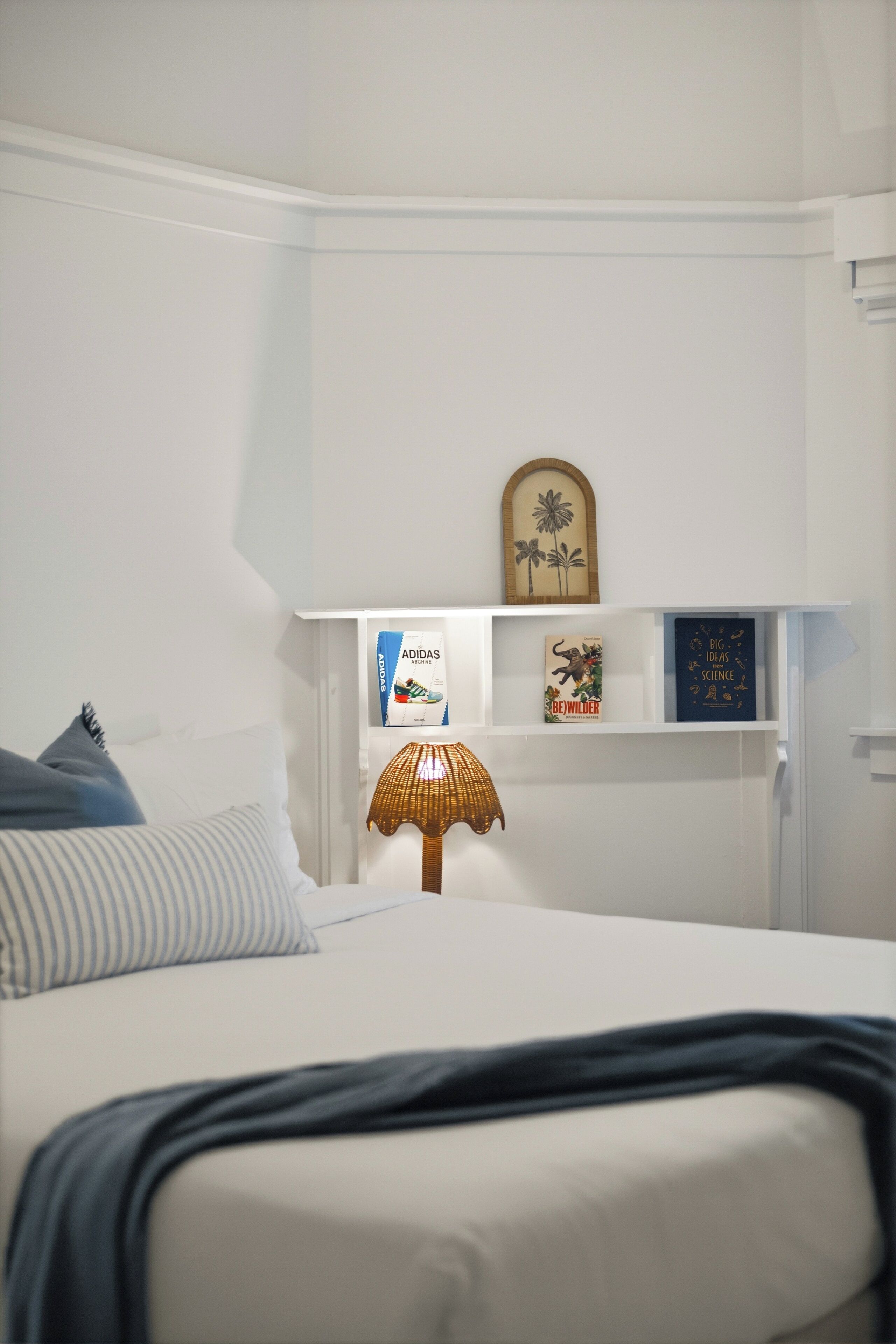 A bedroom with a bed, a bedside lamp, a shelf with books and a framed picture, and a pillow.