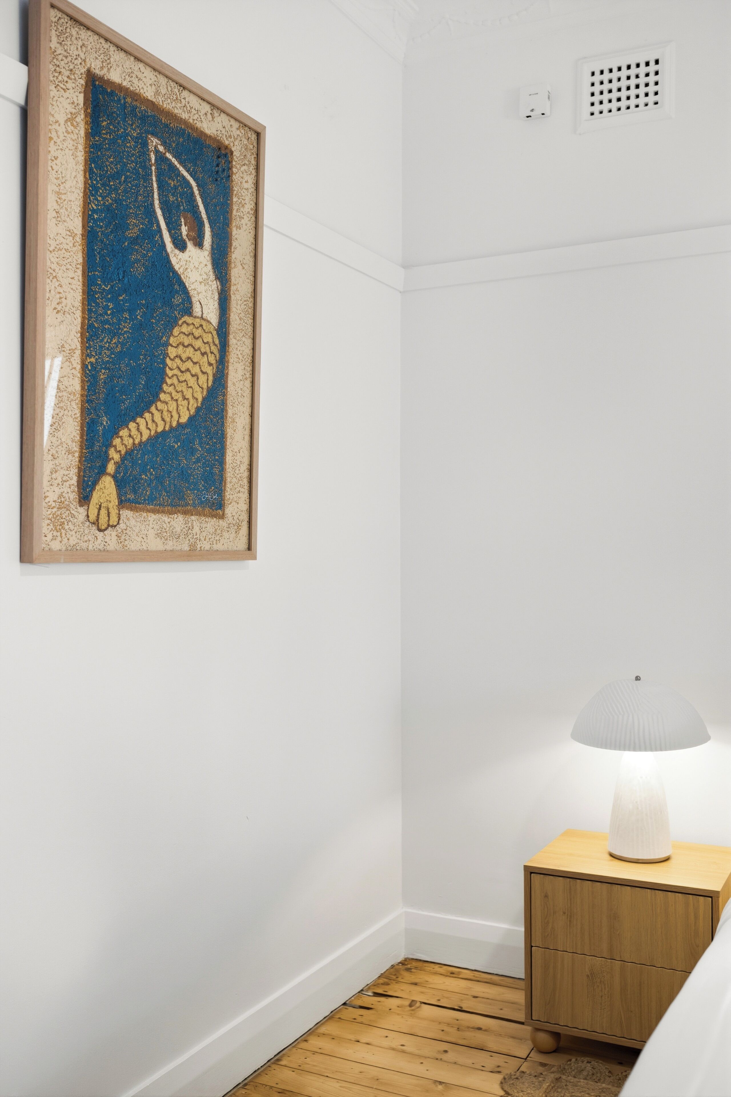 Ein Zimmer mit einem gerahmten Kunstwerk an der Wand, einer Lampe auf einem Nachttisch aus Holz und einem Holzboden.
