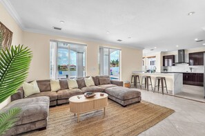 Un salon moderne avec un canapé d’angle, une table basse ronde et une vue sur la cuisine et l’espace extérieur.