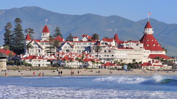 A coastal hotel with multiple red-roofed buildings, a beach in the foreground, and mountains in the background. - Mission Valley Retreat (San Diego)