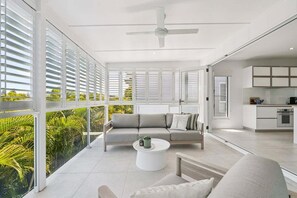 A modern living room with a sofa, coffee table, and a view of greenery through large windows. - The Cheeky Palm - Beachy tropical paradise, perfect for a cheeky cocktail! (Coffs Harbour)