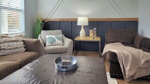 Una sala de estar con un sofá, un sillón y una mesa de centro. Hay una lámpara sobre una mesita auxiliar y una planta en una esquina.