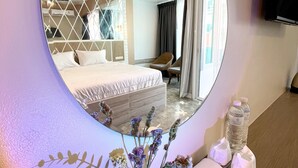 A hotel room with a bed, a chair, a mirror, a vase with flowers, and a tray with toiletries. - Le Cartier Guesthouse (Patong)
