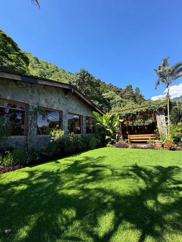 A stone house with large windows, surrounded by lush greenery and a well-maintained lawn. - Guatemala Dreams Grand Chalet (San Lucas Tolimán)