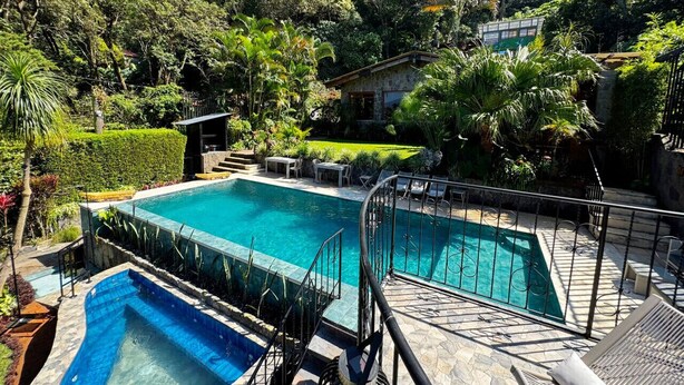 A rectangular swimming pool with a stone deck and metal railings, surrounded by lush greenery and a hillside backdrop. - Guatemala Dreams Grand Chalet (San Lucas Tolimán)