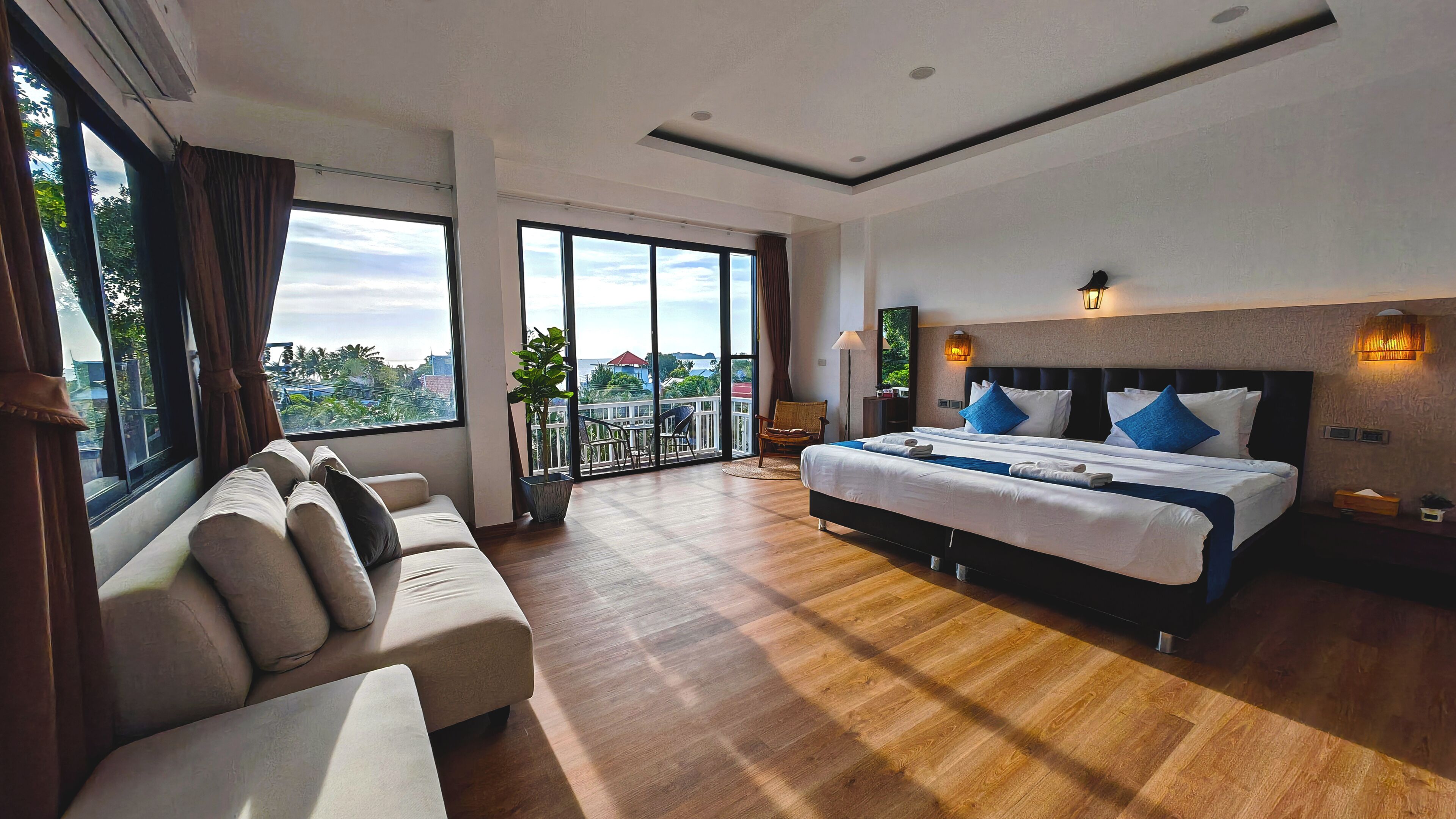 Una habitación de hotel moderna con una cama grande, un sofá, un balcón con vistas y suelo de madera.