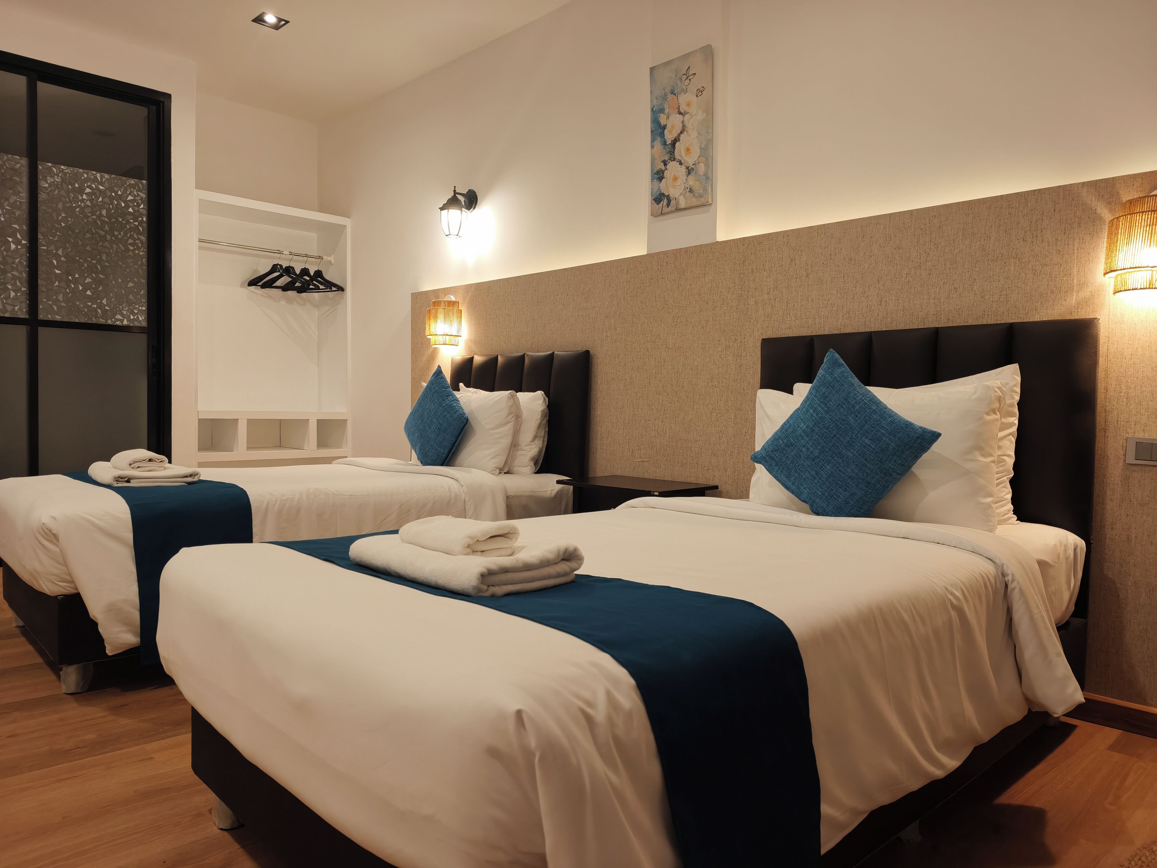 A hotel room with two beds, each with a blue accent pillow, a bedside table, and a wall-mounted lamp.
