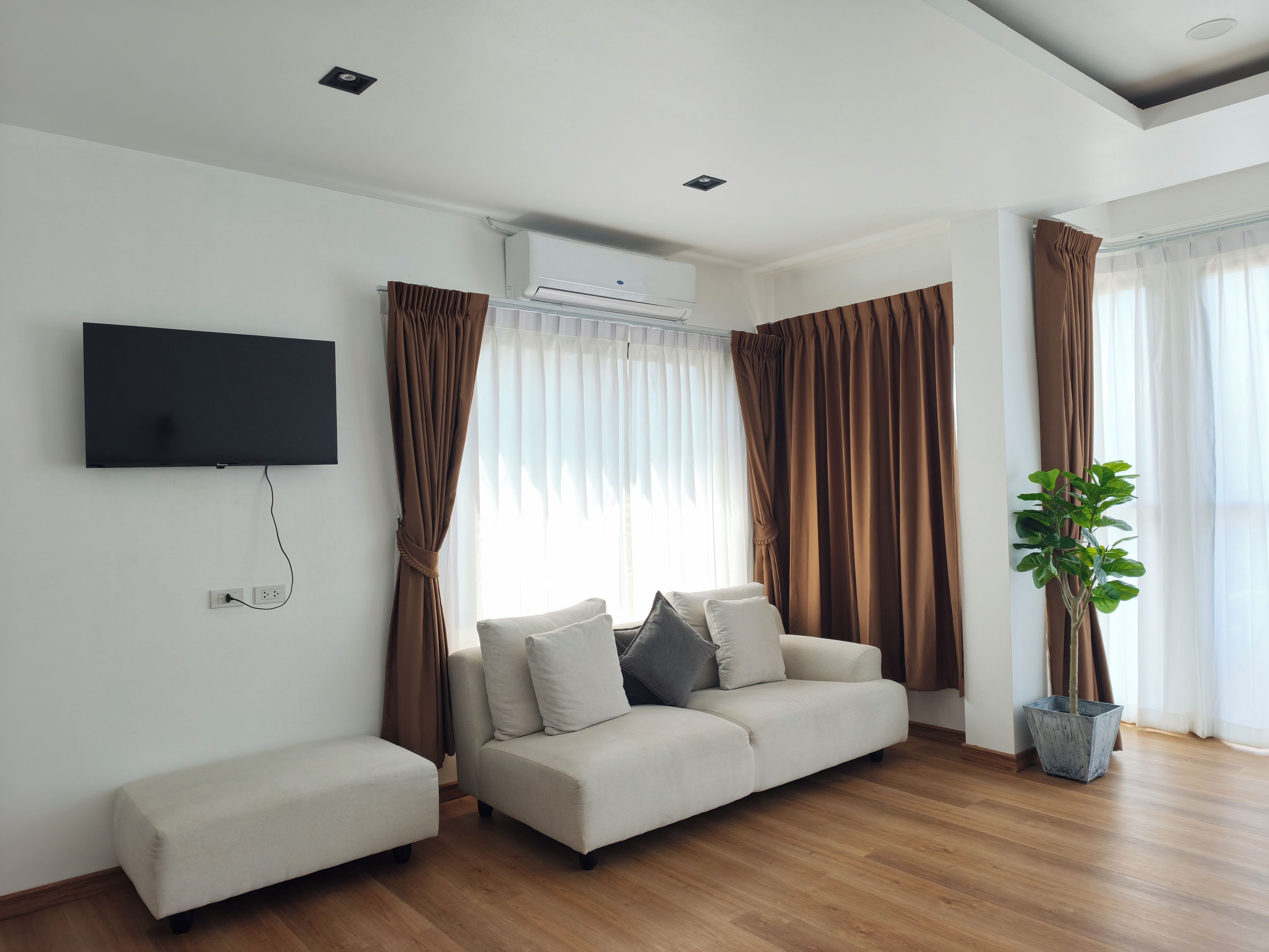 Una sala de estar moderna con un televisor de pantalla plana montado en la pared, un sofá color beige con cojines, un pequeño banco y una planta en maceta.