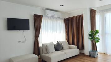 Una sala de estar moderna con un televisor de pantalla plana montado en la pared, un sofá color beige con cojines, un pequeño banco y una planta en maceta.