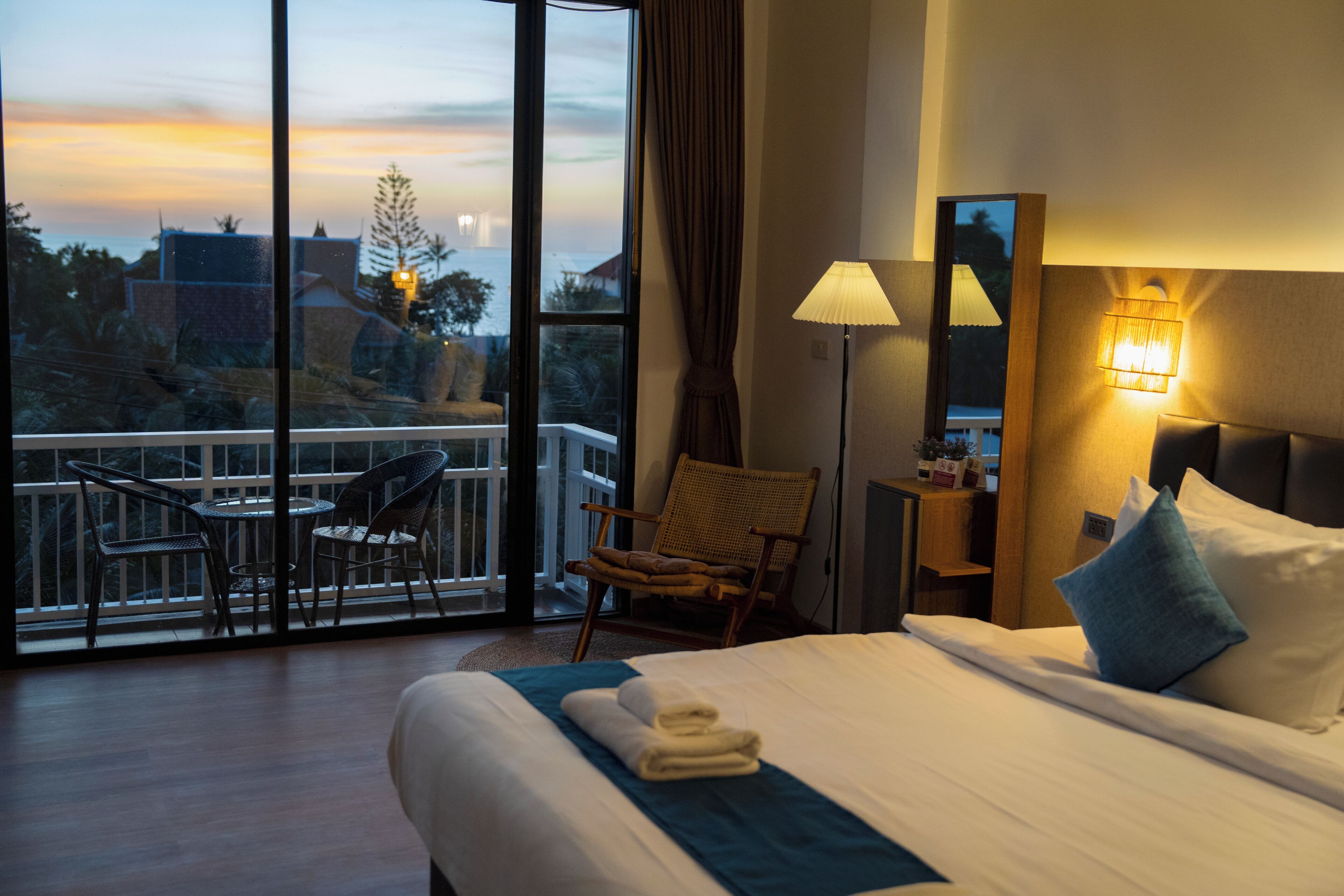 A hotel room with a balcony, a bed with pillows, a chair, a table, and a lamp.