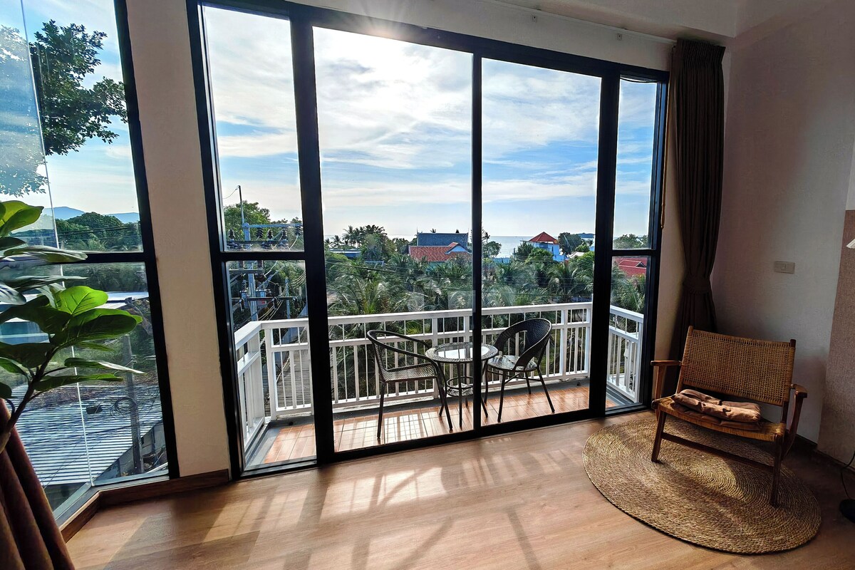 A room with a large sliding glass door leading to a balcony with a view of the sea and buildings. There is a wicker chair and a floor lamp on the balcony.