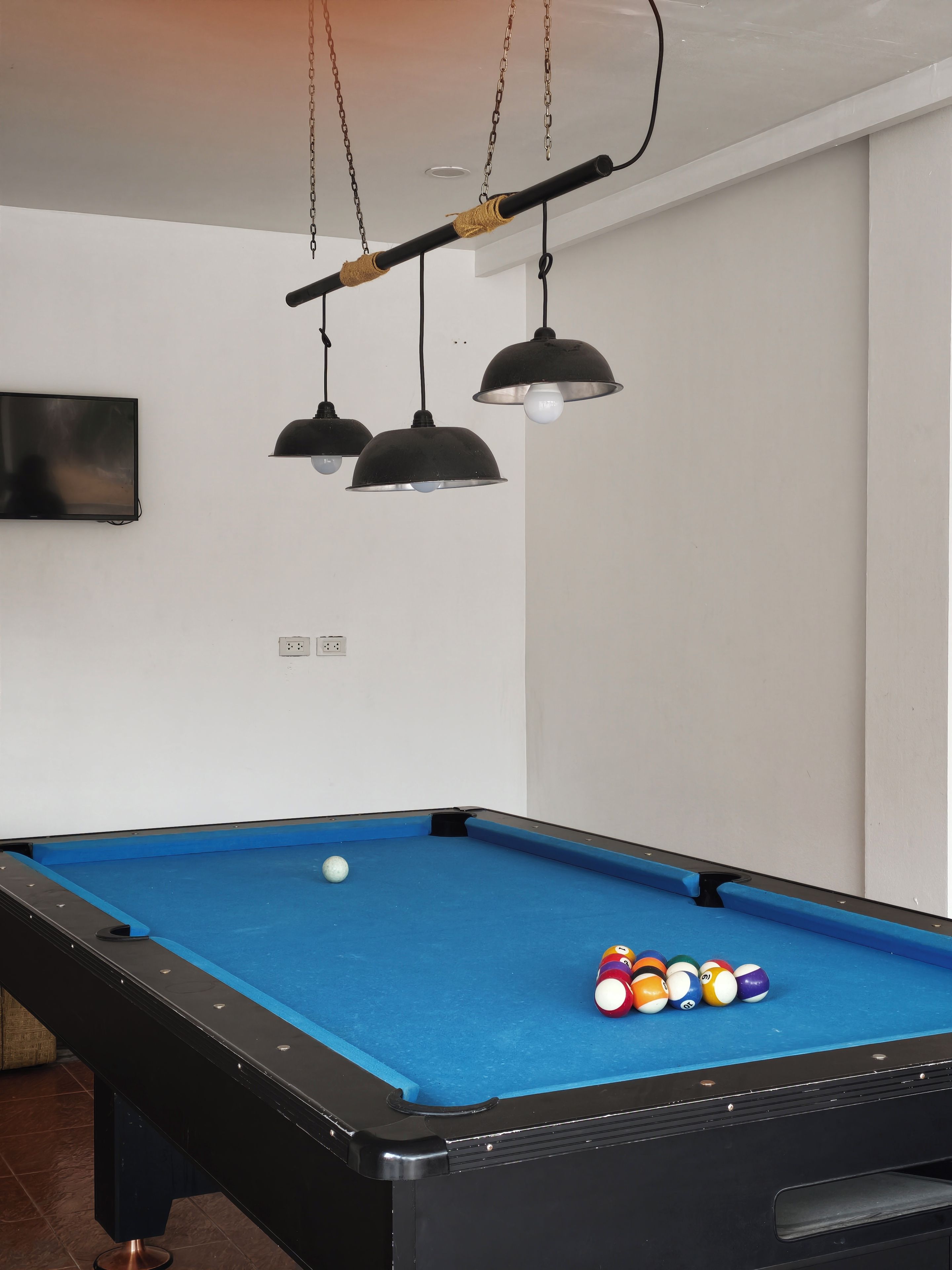 Una mesa de billar con bolas, un televisor y lámparas colgantes en una habitación con paredes blancas.
