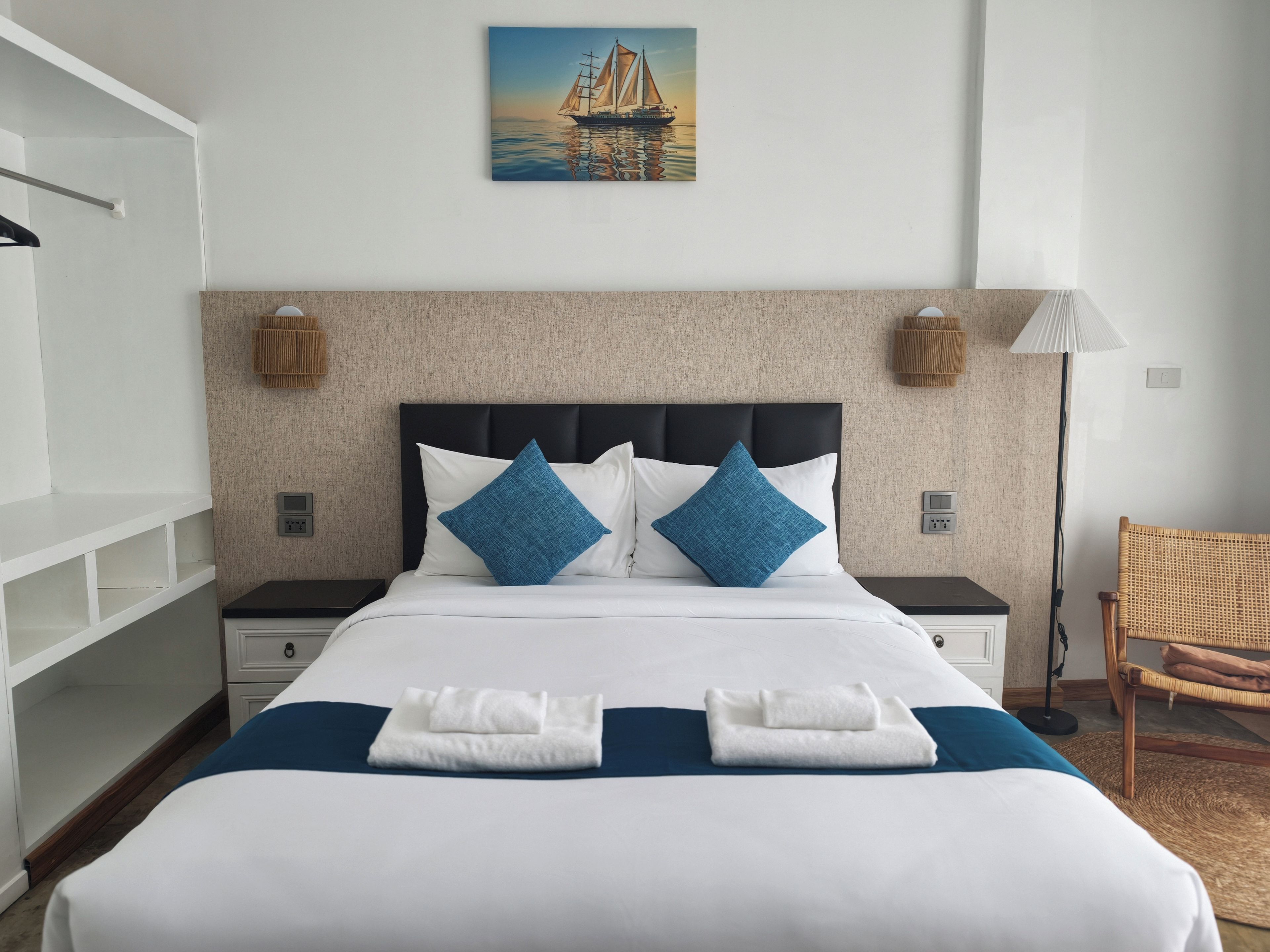 Habitación de hotel con cama, dos mesitas de noche, una lámpara de pared, un sillón de mimbre y un cuadro de un velero.