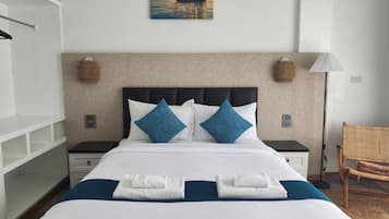 Habitación de hotel con cama, dos mesitas de noche, una lámpara de pared, un sillón de mimbre y un cuadro de un velero.