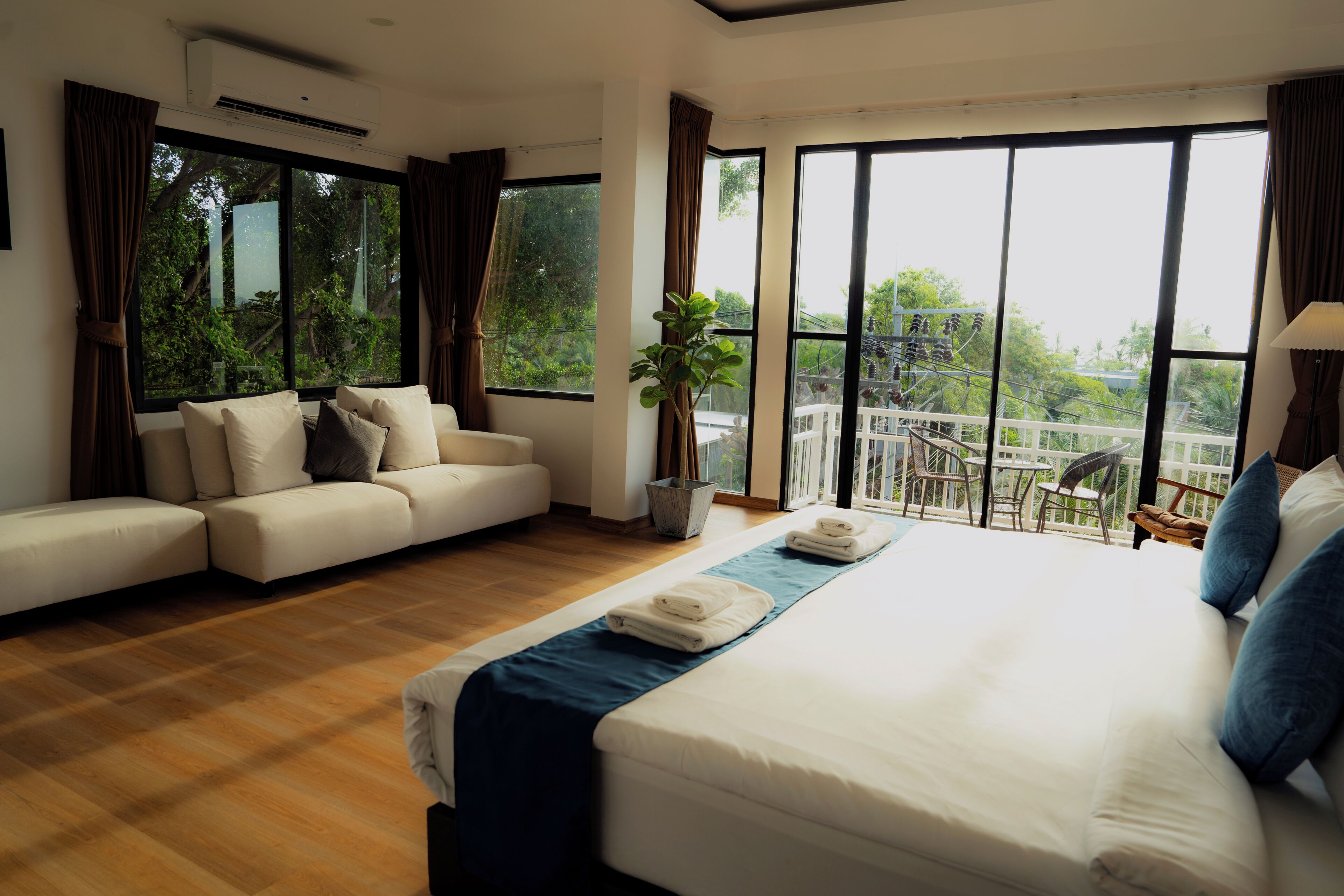 A modern hotel room with a large bed, a sofa, and a balcony with a view of greenery.