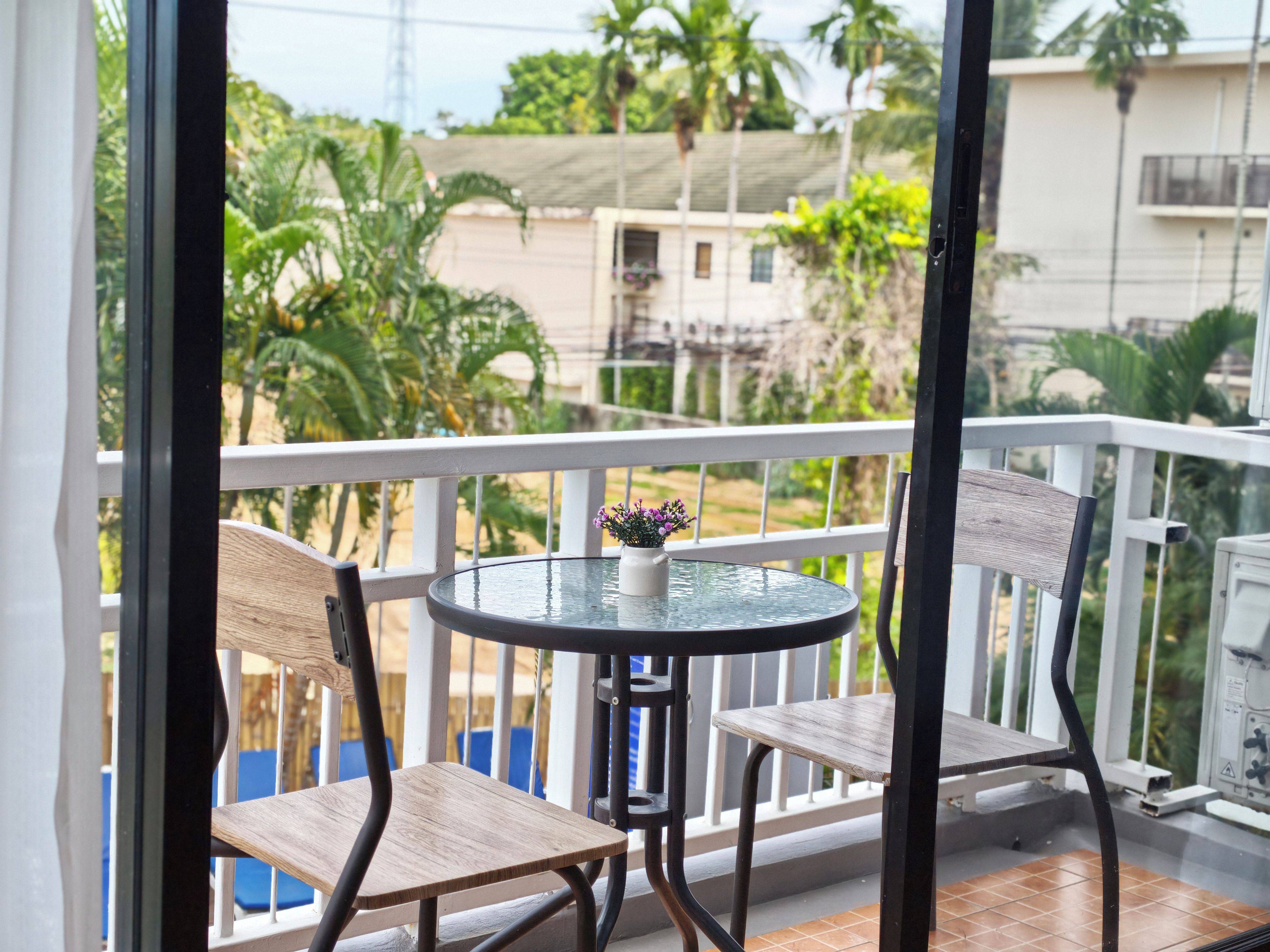 Un balcon avec une table ronde et deux chaises, un pot de fleurs et une vue sur la verdure et les bâtiments.