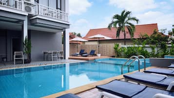 Una piscina con sillones de descanso, un área de estar techada y un edificio con unidades de aire acondicionado.