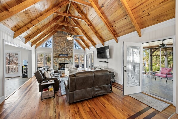 A spacious living room with a stone fireplace, wooden ceiling beams, a large leather sofa, and a television mounted on the wall.