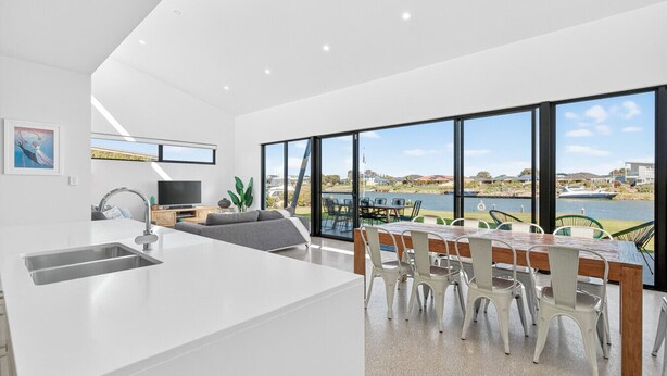 A modern kitchen with a large island, dining area with white chairs, and large windows overlooking a body of water.