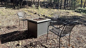 Ein Sitzbereich im Freien mit zwei Metallstühlen und einem quadratischen Feuerkorb, eingebettet in eine bewaldete Landschaft.