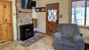 Ein Wohnzimmer mit Kamin aus Stein, einem darüber hängenden Fernseher, einer Holztür und einem Fenster mit Jalousien.