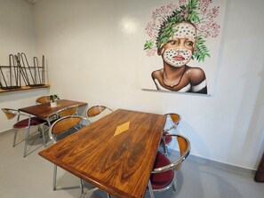 A dining area with wooden tables and red cushioned chairs, a wall-mounted painting, and metal chairs on a shelf.